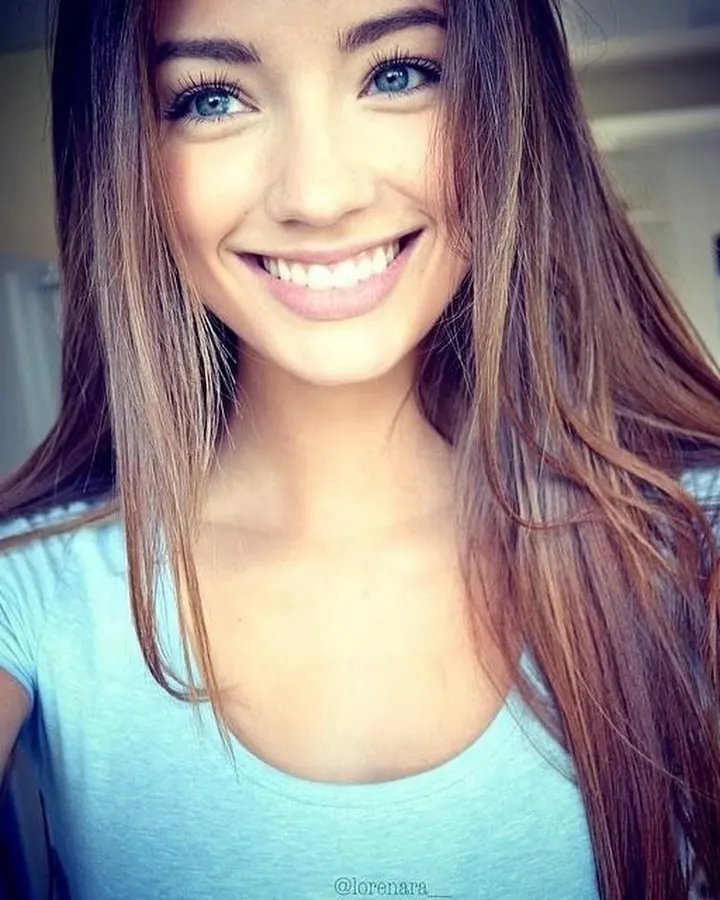 Jeune femme aux cheveux bruns souriant largement à l'appareil photo dans un cadre intérieur.