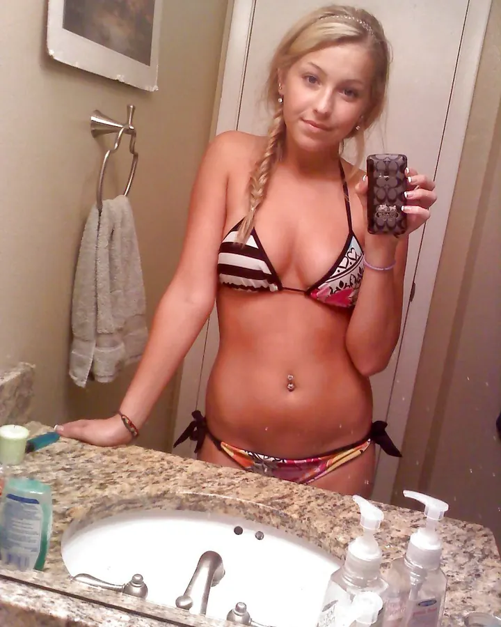 Jeune femme blonde en maillot de bain multicolore prenant un selfie dans une salle de bain avec un miroir et un lavabo.