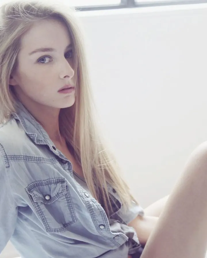 Jeune femme blonde aux cheveux longs, portant une chemise en jean clair, assise sur un lit blanc dans une pièce lumineuse.