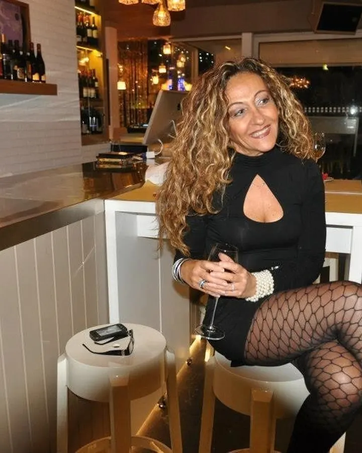 Femme blonde en tenue noire, assise dans un bar, tenant un verre de vin