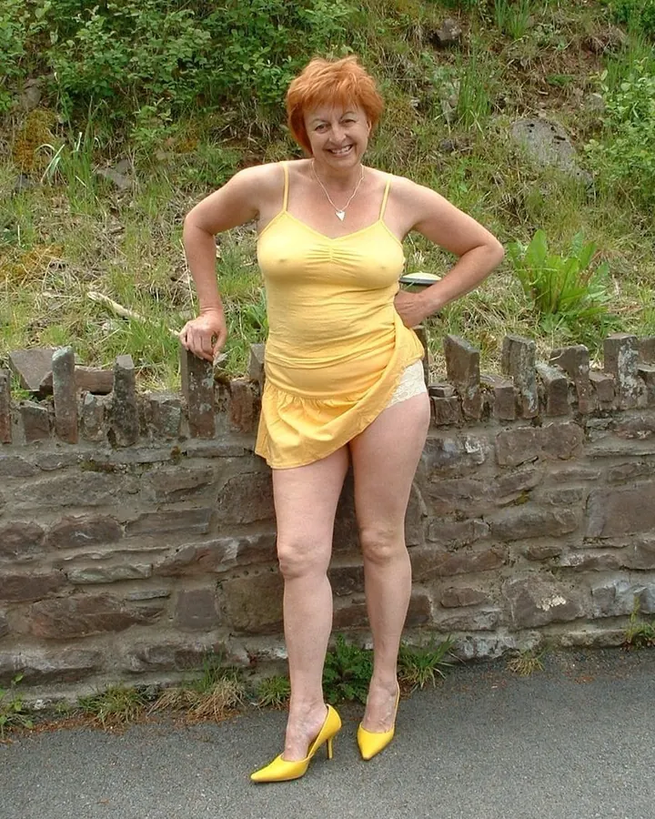 Femme plus âgée aux cheveux roux souriante, debout devant un mur en pierre, vêtue d'une robe jaune et de chaussures jaunes.