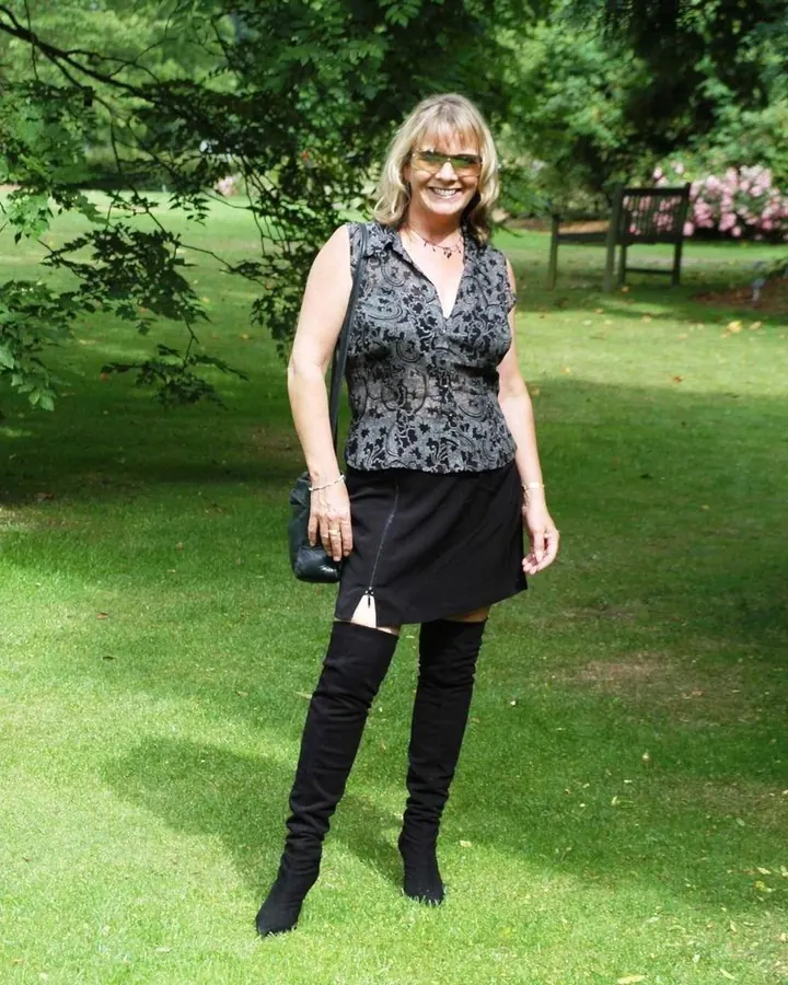 Femme aux cheveux blonds platine, lunettes de soleil, souriante, debout dans un parc en jupe noire et bottes hautes.