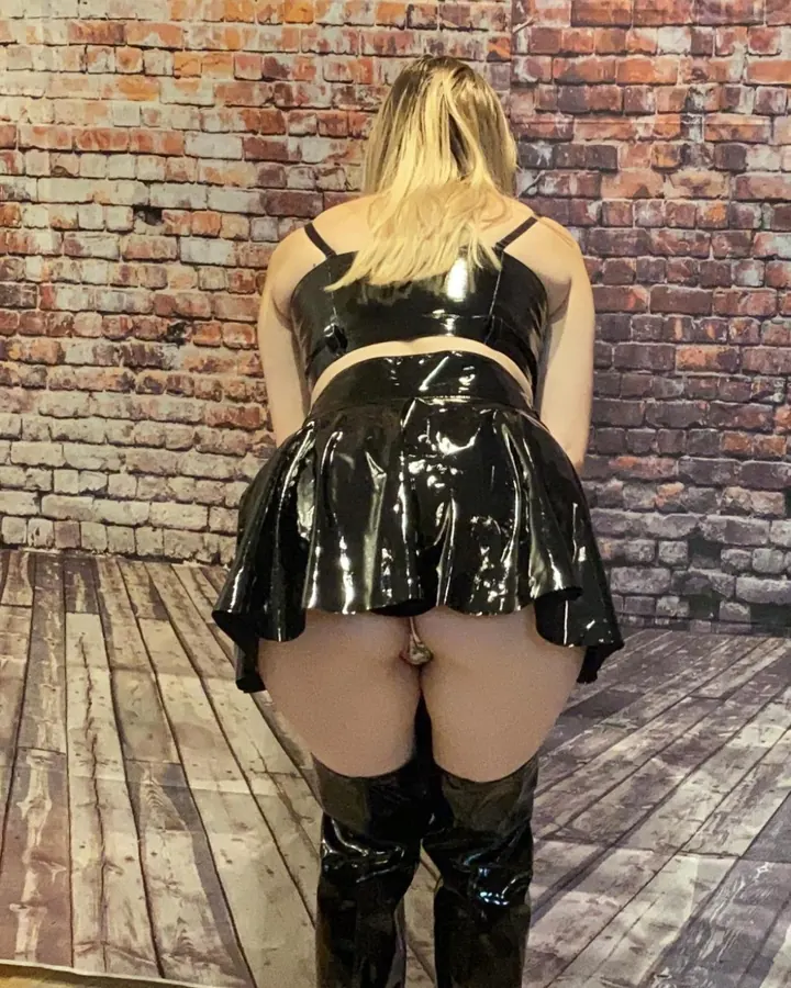 Femme blonde vue de dos, portant une tenue en latex noir, basques, bottes hautes, devant un mur de briques.