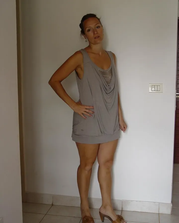 Femme mûre en robe grise, mains sur les hanches, regard direct, ambiance intime