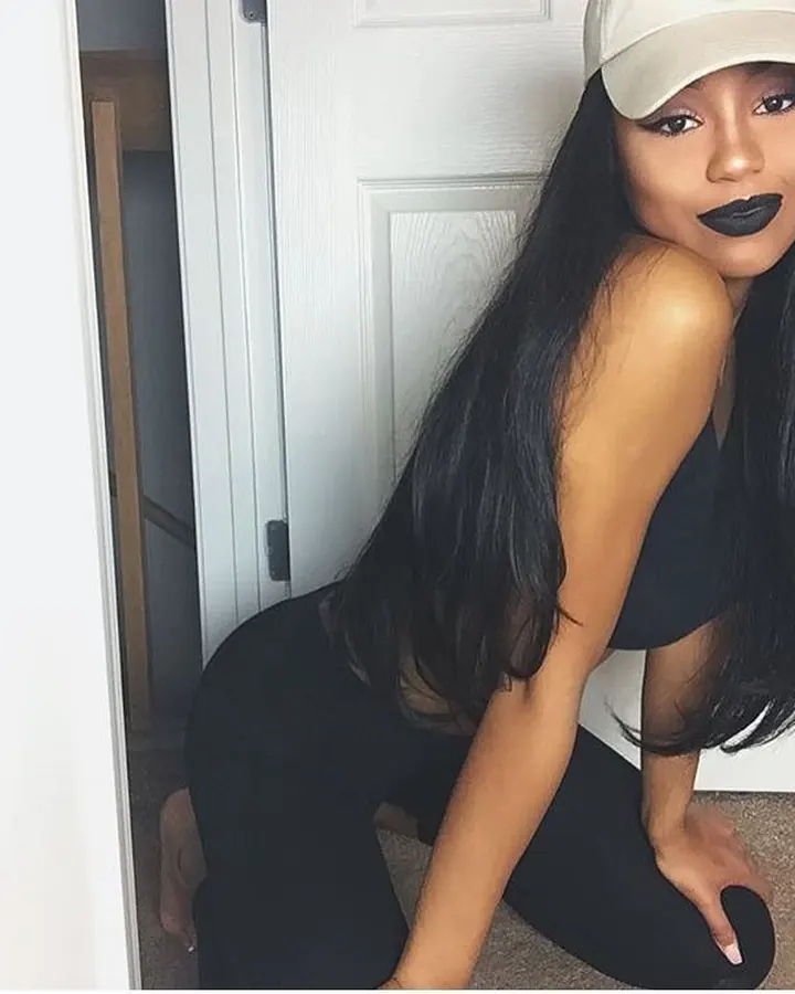 Femme aux cheveux longs noirs, portant un body noir, une casquette blanche et un maquillage sombre, posant près d'une porte blanche