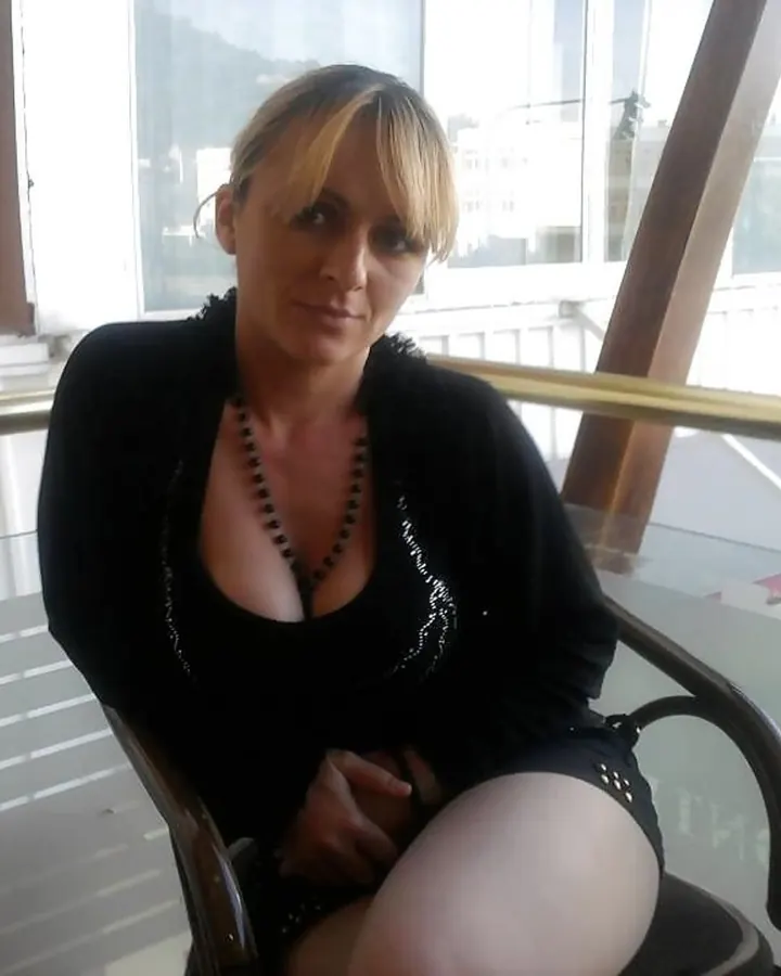 Femme blonde portant un haut noir, assise sur une chaise dans un espace lumineux avec une rambarde métallique.