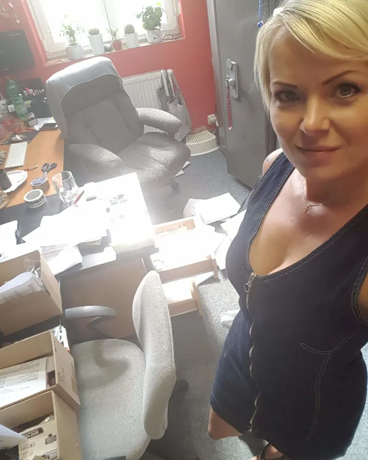 Femme blonde portant un haut noir, assise dans un bureau encombré avec des boîtes et un fauteuil de jeu.