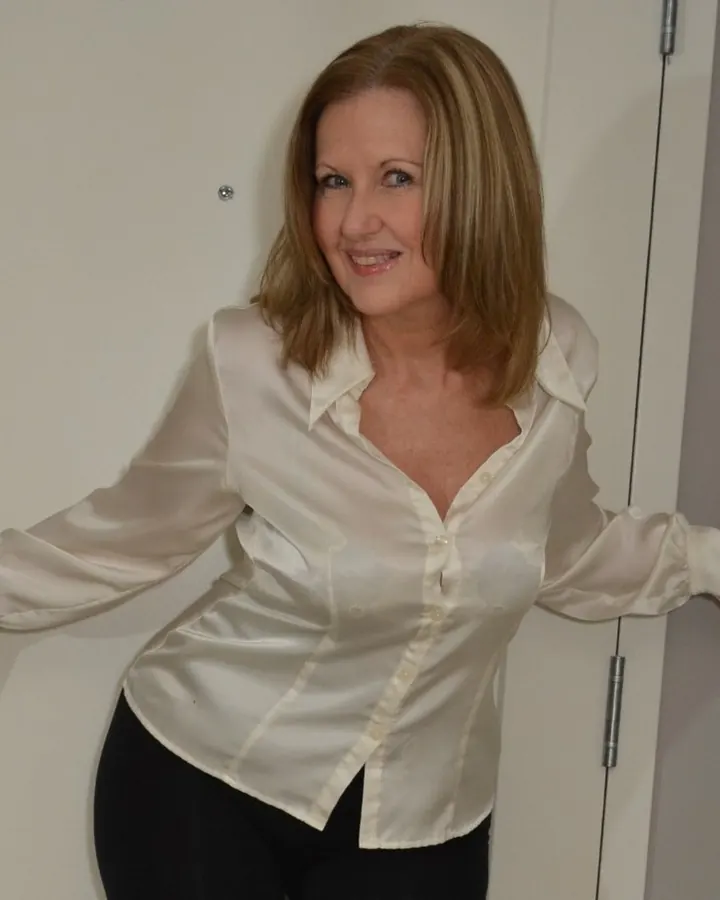 Femme aux cheveux blonds, portant un haut noir et une jupe noire, allongée sur un lit blanc avec un sourire.