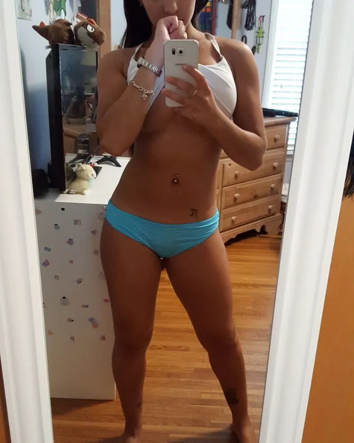 Femme prenant un selfie devant un miroir, vêtue d’un haut blanc et d’un string bleu, dans une chambre.