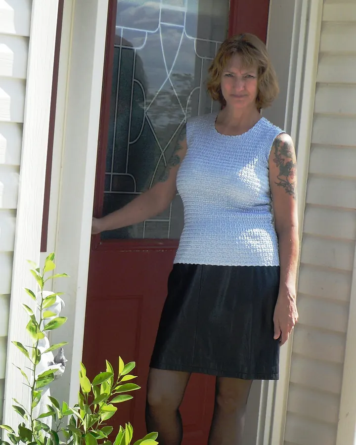 Femme blonde en tenue décontractée, debout devant une porte extérieure, avec des tatouages aux bras