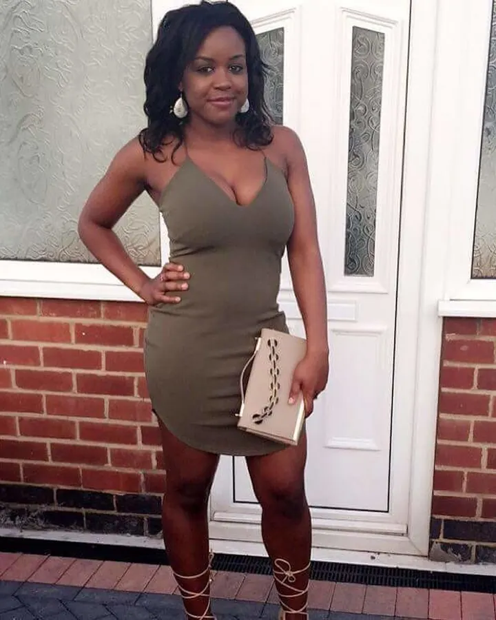 Femme africaine aux cheveux bruns-noirs, souriante, portant une robe verte ajustée, tenant un petit sac beige, debout devant une porte blanche et un mur de briques.