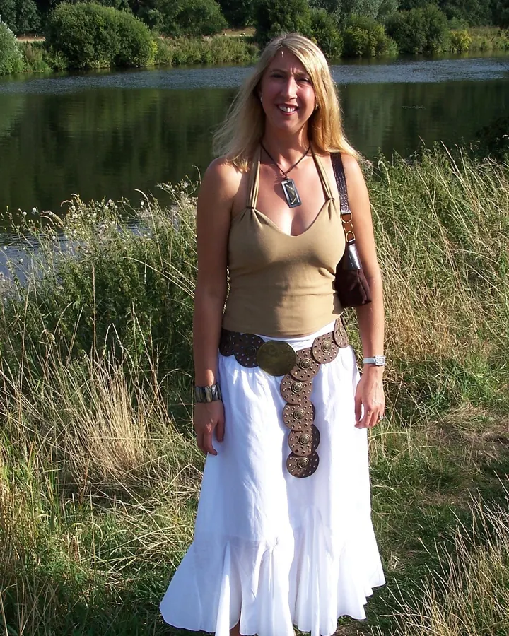 Femme aux cheveux blonds, vêtue d'une robe noire, assise sur une chaise en bois, les jambes écartées