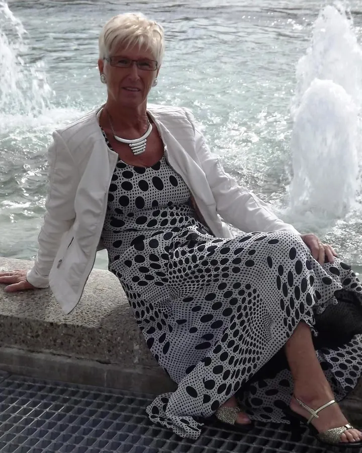 Femme aux cheveux blancs en pull bleu clair, lunettes de soleil, collier avec médaille, assise contre mur briques