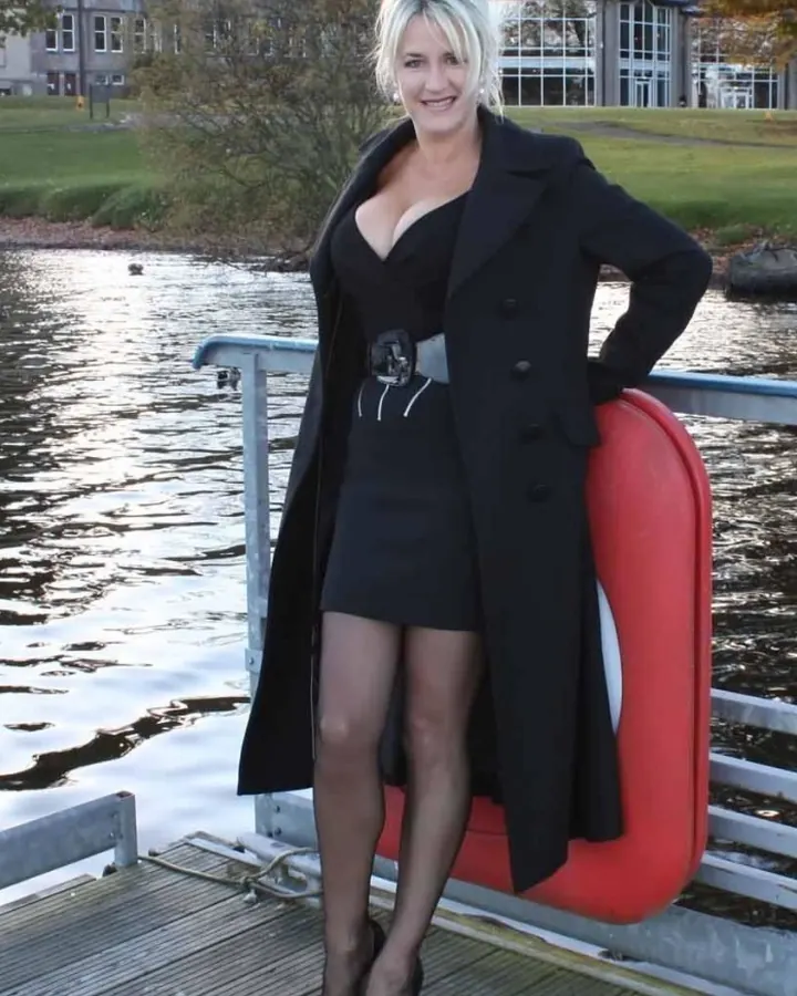 Femme aux cheveux platine portant une robe noire et un manteau noir, debout sur un pont en bois près d'un lac, souriante.