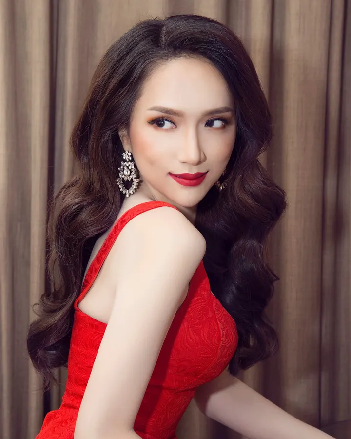 Femme aux cheveux châtains portant une robe rouge, souriant avec des boucles d'oreilles élégantes, devant un rideau beige.