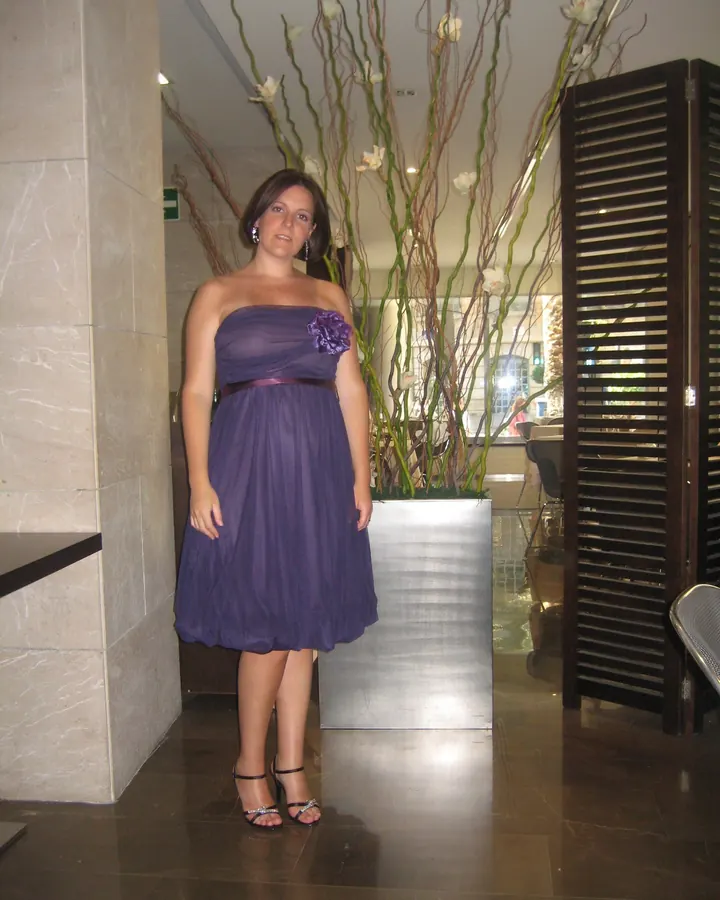 Femme en robe violette sans bretelles, debout dans un hall élégant avec des plantes en arrière-plan.