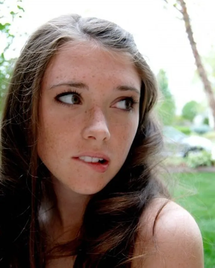 Jeune femme aux cheveux bruns, en extérieur, regardant de côté avec une expression curieuse.