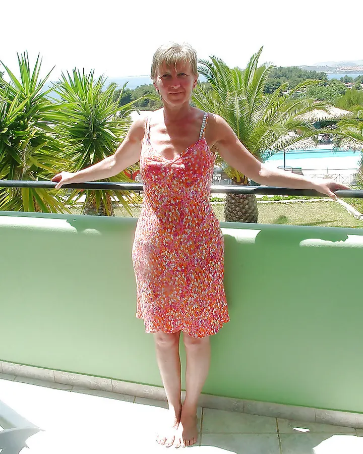 Femme aux cheveux blonds courts, souriante, debout sur une terrasse, portant une robe fleurie rose et orange.